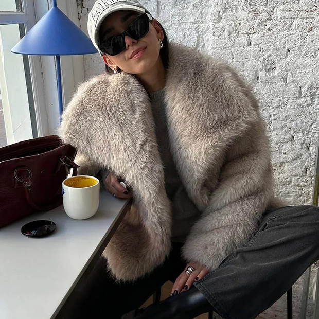 Person wearing a fluffy beige coat sitting at a table with a cup and handbag.