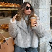 Woman in a fluffy gray coat holding a coffee cup, standing against a stone wall with pastries in the background.