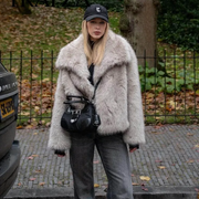 Person wearing a fluffy gray coat and black cap in an outdoor setting with a car and fence in the background.